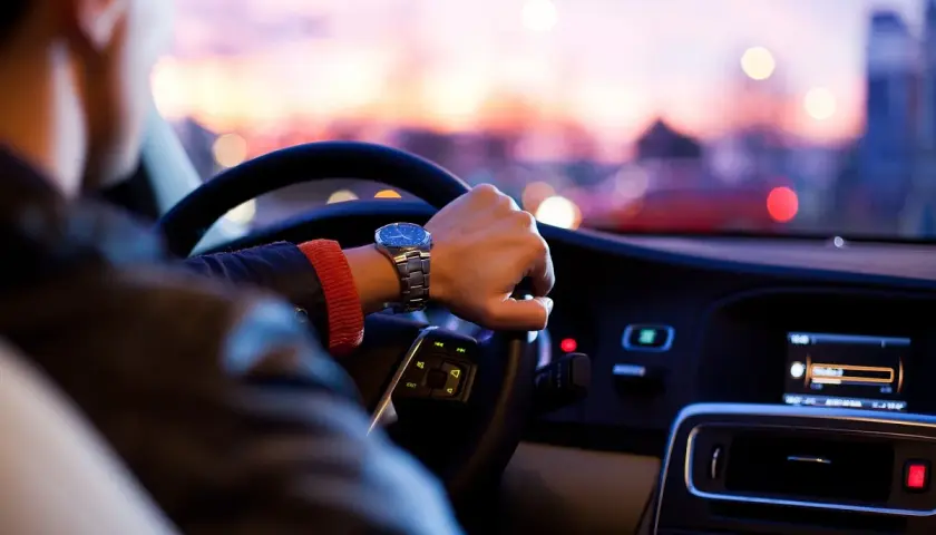 Hombre con reloj conduce a mayor velocidad, con luces de ciudad borrosas al atardecer.