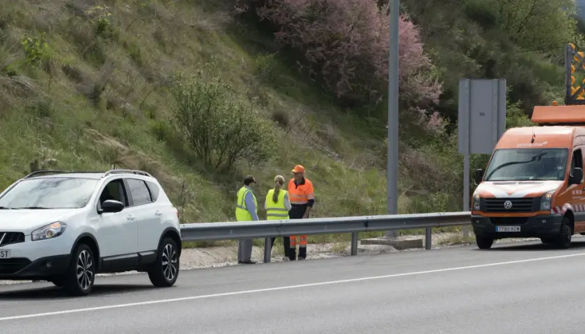 Personal de autopistas atiende un incidente en la carretera. Un coche blanco y una furgoneta naranja con señalización de emergencia están detenidos.