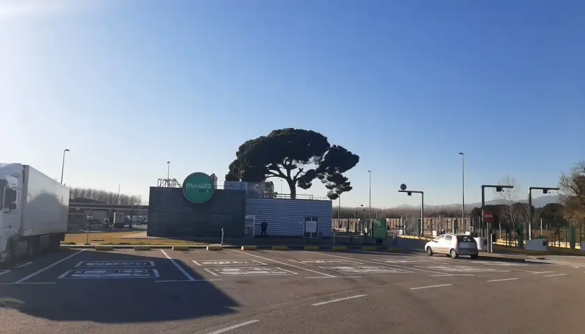 Un área de parking camiones con un gran pino y el logo de "Mawiz Truck". Un camión blanco está aparcado a la izquierda.
