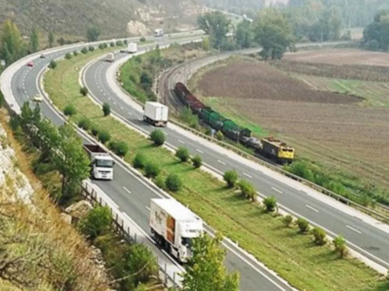 Autopista con camiones y tren de carga. Un viaje escénico de Burgos a Zaragoza sin peaje, con paisajes verdes y campos.
