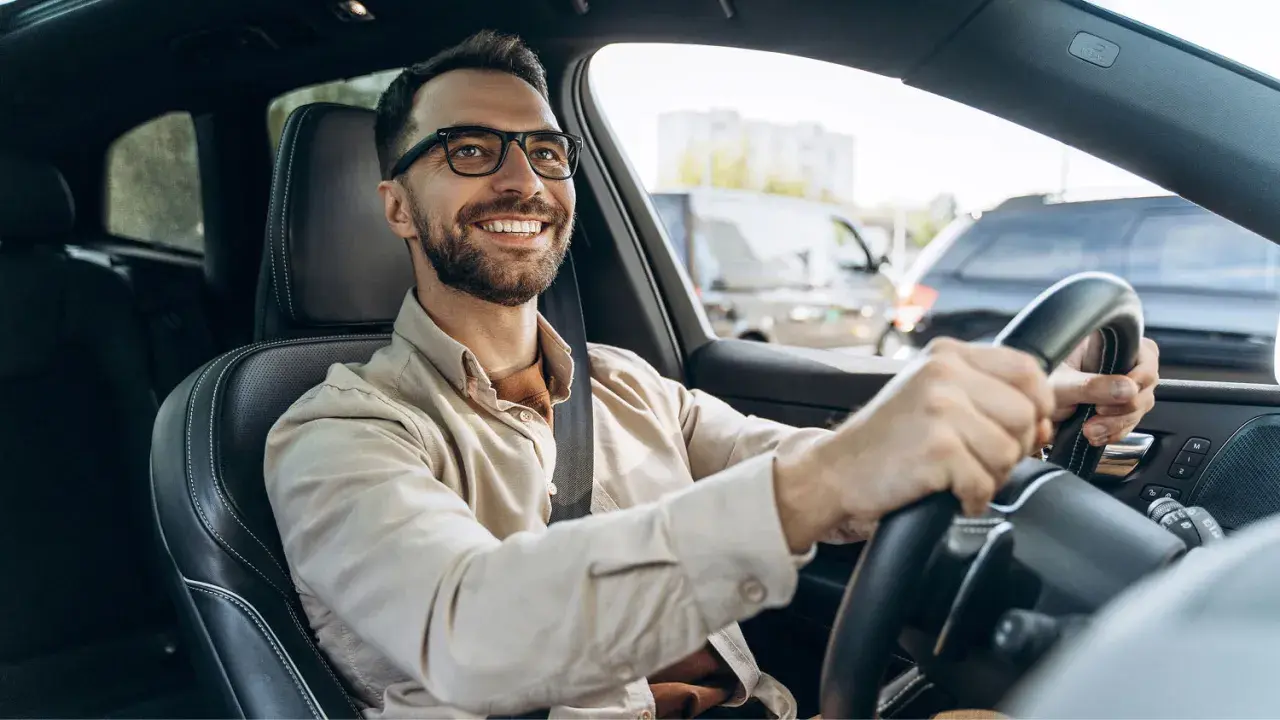 Hombre sonriente al volante, con gafas y camisa clara. Viaja en coche, quizás de camino a una cita importante, con la tranquilidad que ofrece la seguridad de Unicaja.
