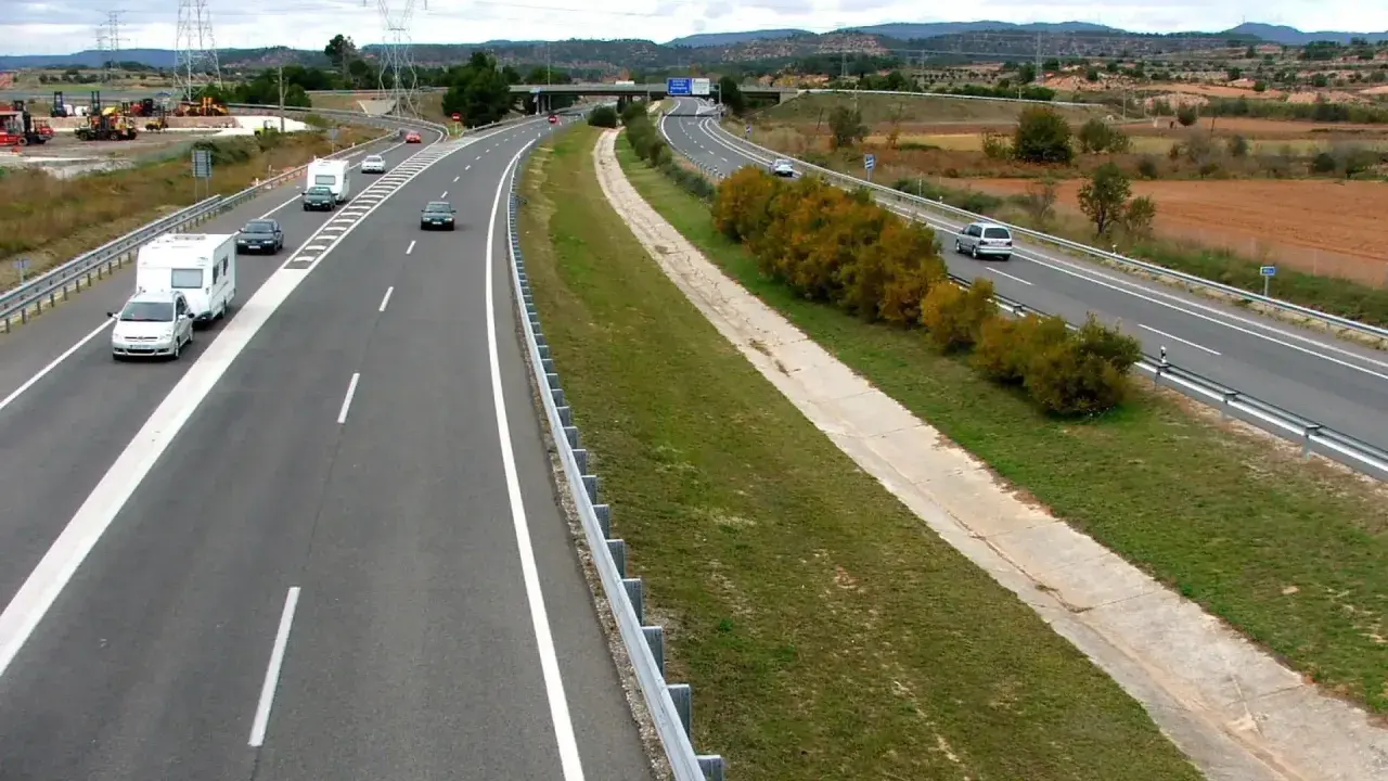 Autopista con varios coches y caravanas. Se aprecian las áreas de servicio AP2 al fondo, con vegetación y campos.