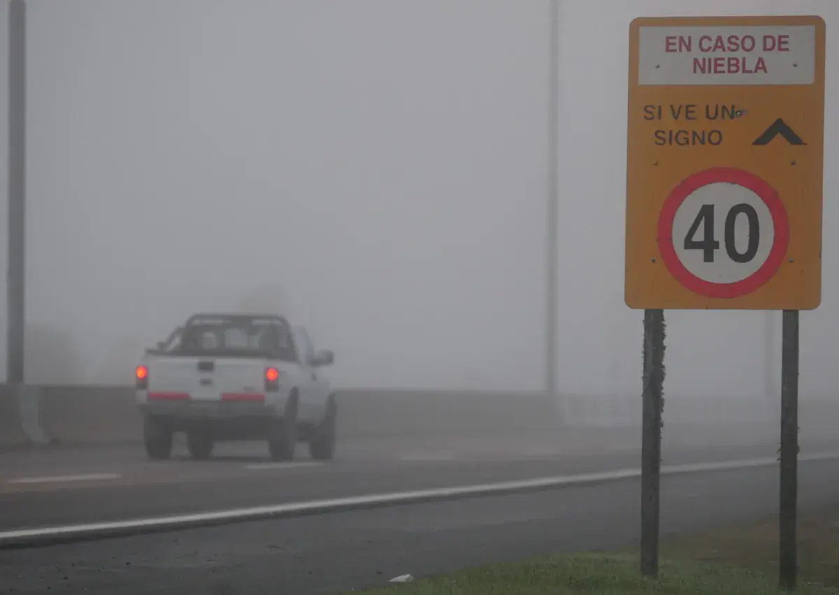 Camioneta blanca avanza en **niebla densa** bajo un **alumbrado** de señalización vial que indica 40 km/h.