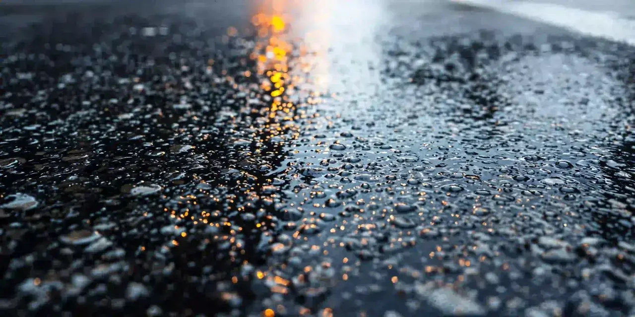 Gotas de agua brillan sobre la carretera mojada, reflejando luces anaranjadas.