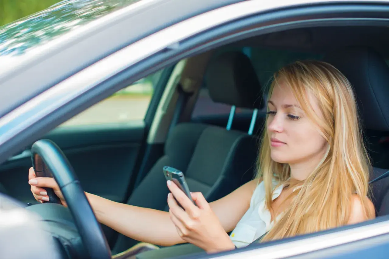Mujer rubia al volante, mirando su móvil. Es importante evitar distracciones al conducir para mayor seguridad.