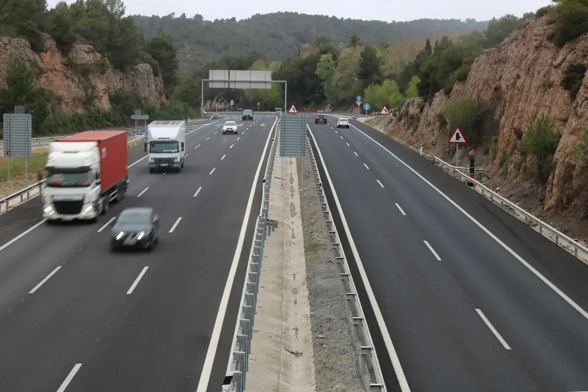 Tráfico en las autopistes catalanes, con camiones y coches circulando por una carretera de varios carriles rodeada de vegetación y rocas.