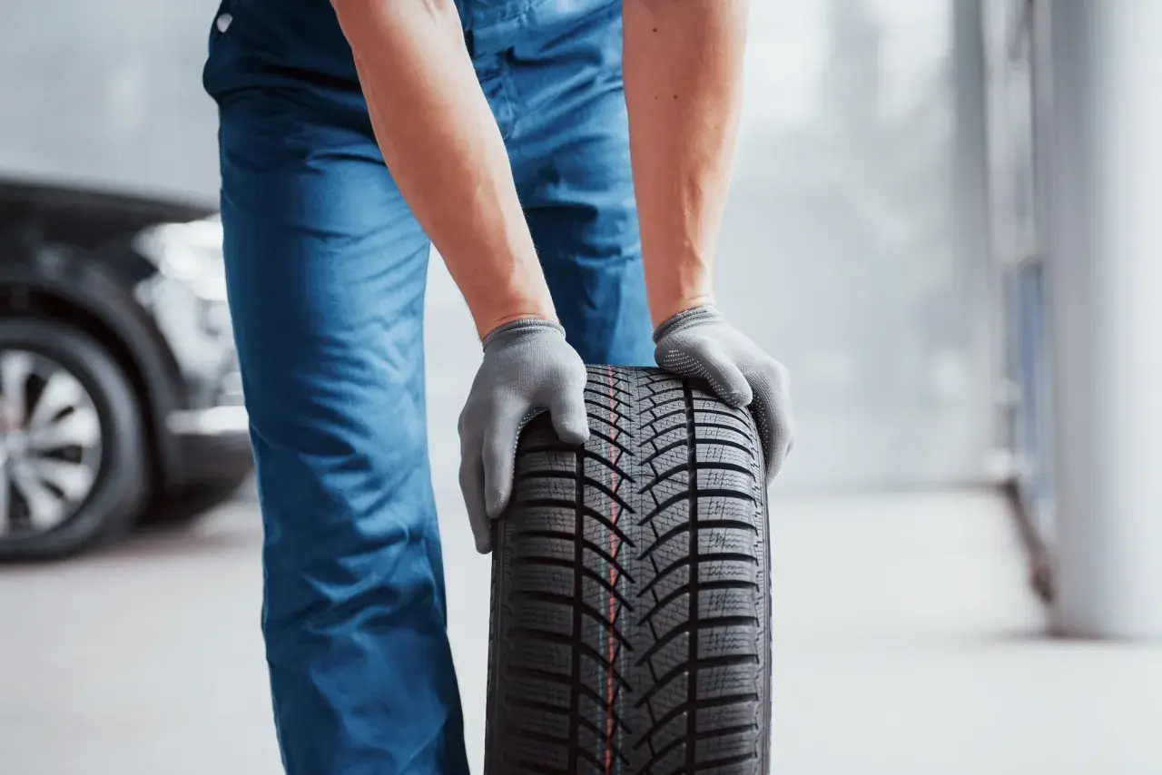 Man con guantes grises sostiene un neumático nuevo, listo para mejorar la adherencia del coche.