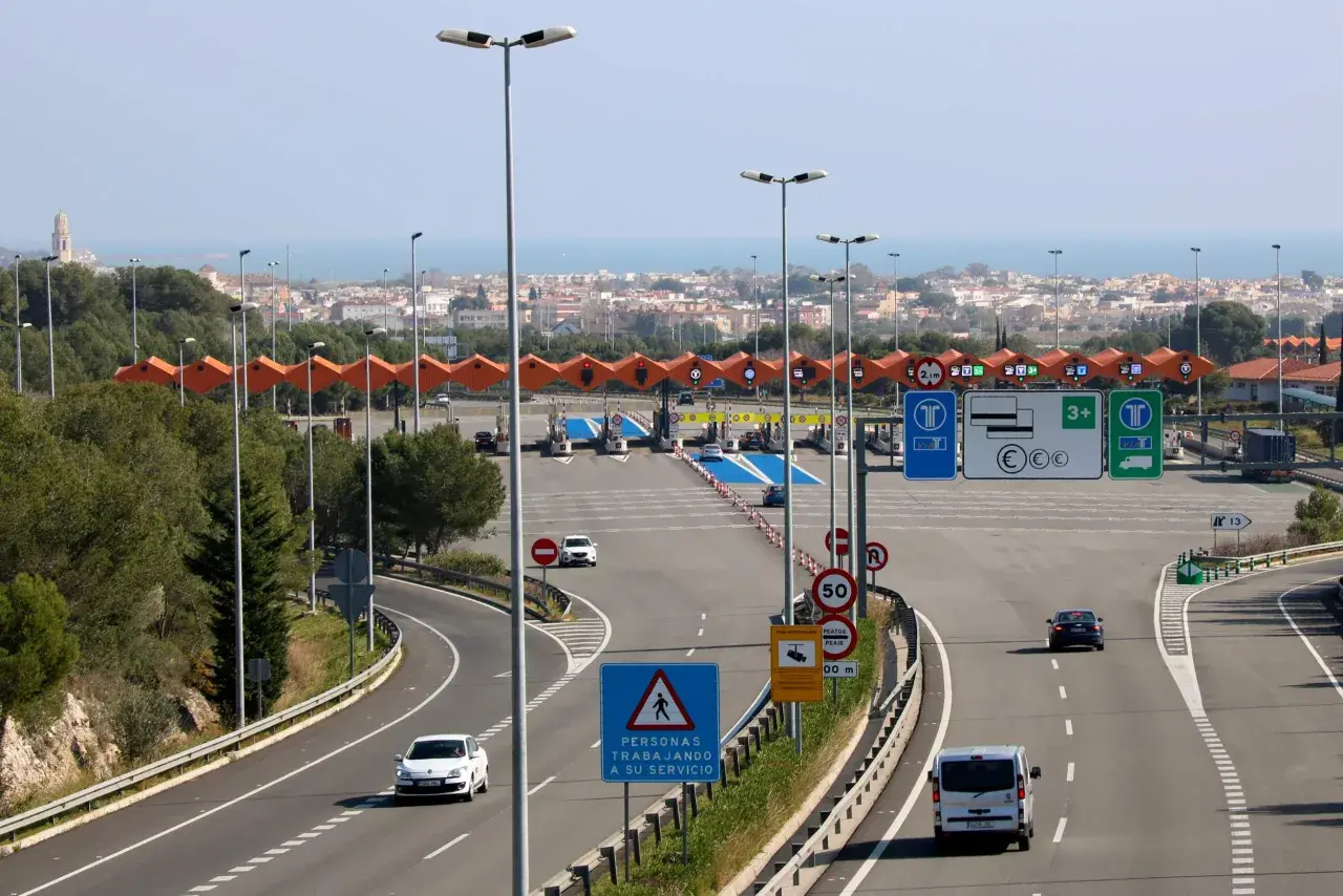 Peaje en la autopista Pau Casals con varios carriles, señales de tráfico y vehículos circulando. Al fondo, un paisaje urbano y el mar.