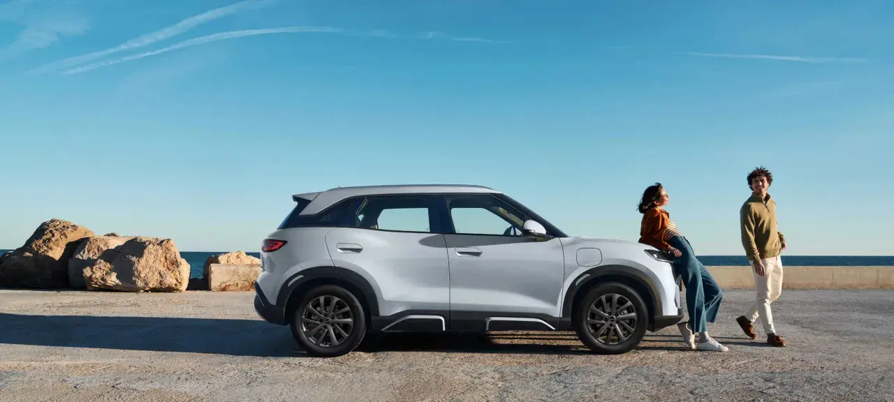 Pareja junto a un moderno coche híbrido blanco, con el mar y rocas al fondo.