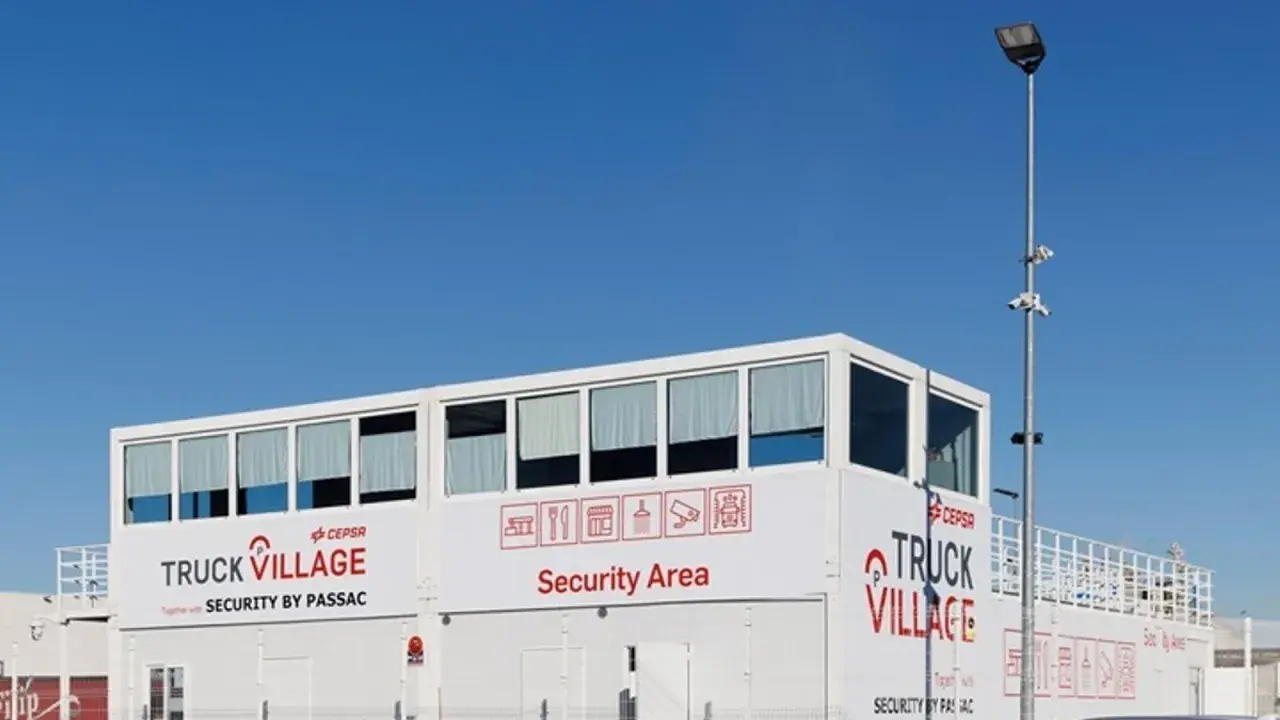 Edificio blanco con letreros "TRUCK VILLAGE" y "Security Area". Un poste con cámaras y luz ilumina el aparcamiento de camiones.
