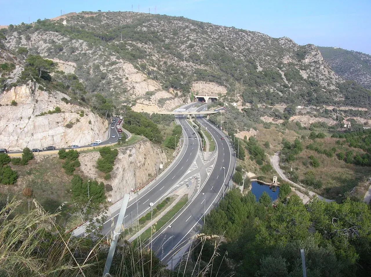 La autopista C32 serpentea entre montañas rocosas y vegetación, con un túnel y un puente.