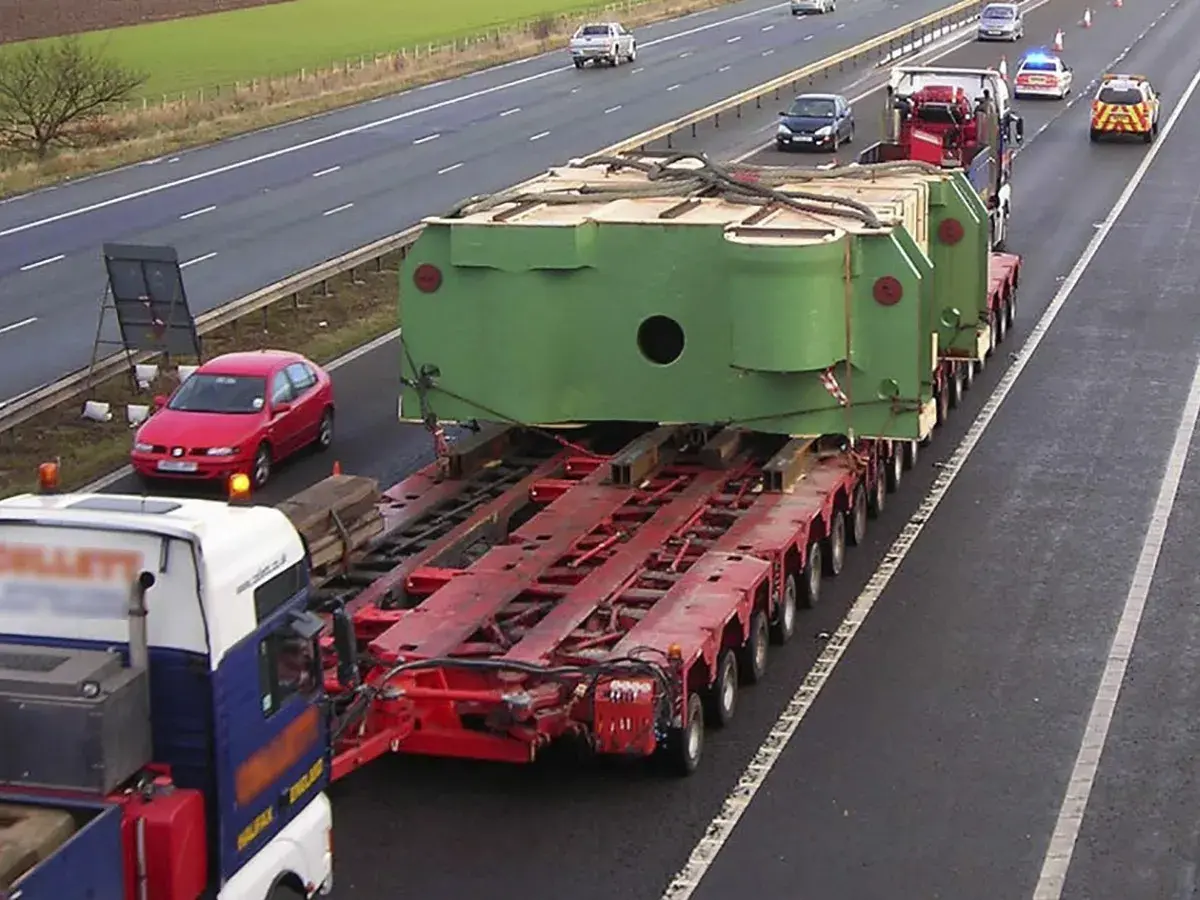 Un camión transporta una enorme pieza verde sobre una plataforma de múltiples ruedas. Un coche rojo y vehículos policiales acompañan este convoy de transportes especiales en la autopista.