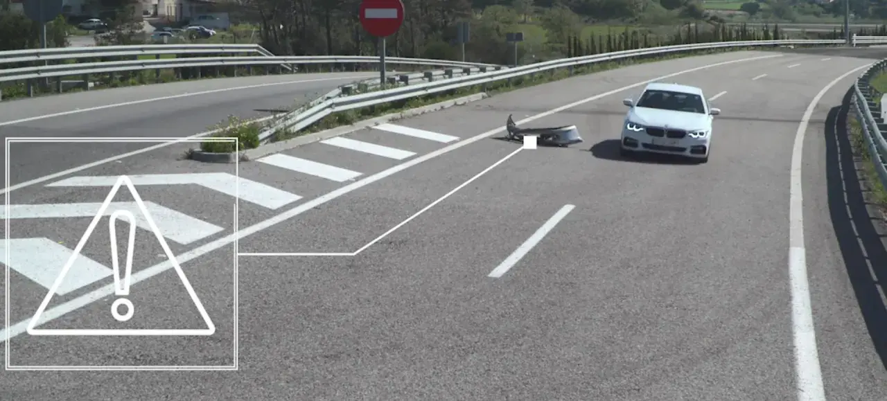 Un coche blanco se detiene cerca de un parachoques dañado en la carretera, indicando un accidente por objeto en la carretera.