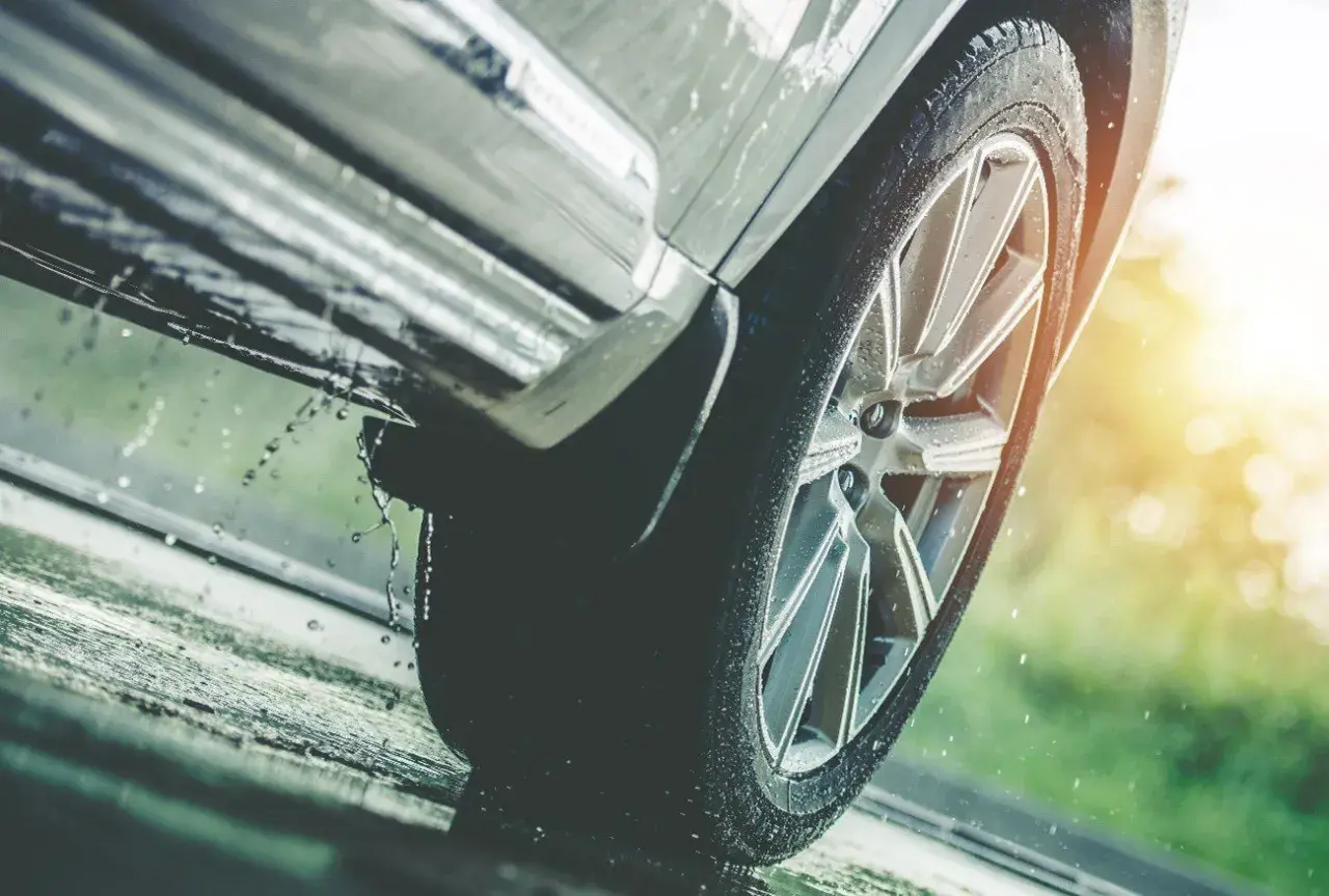 Rueda de coche sobre asfalto mojado, con gotas de agua salpicando, indicando una posible **pérdida de adherencia** en condiciones de lluvia.