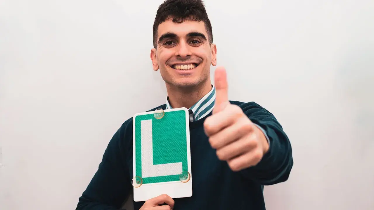 Joven sonriente con letrero "L" y pulgar arriba, listo para ser novel al volante.