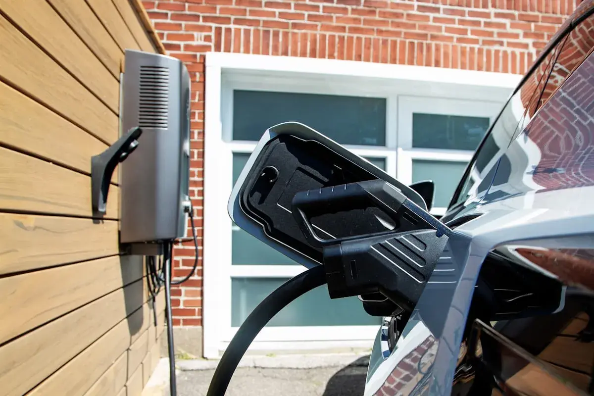 Cargador bidireccional V2H conectado a un coche eléctrico. La unidad de carga está montada en una pared de madera, con una casa de ladrillo de fondo.
