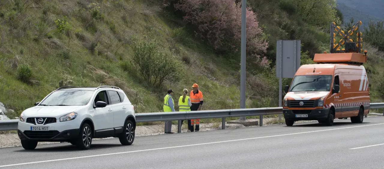 Personal de autopistas atiende un incidente en la carretera. Un coche blanco y una furgoneta naranja con señalización de emergencia están detenidos.