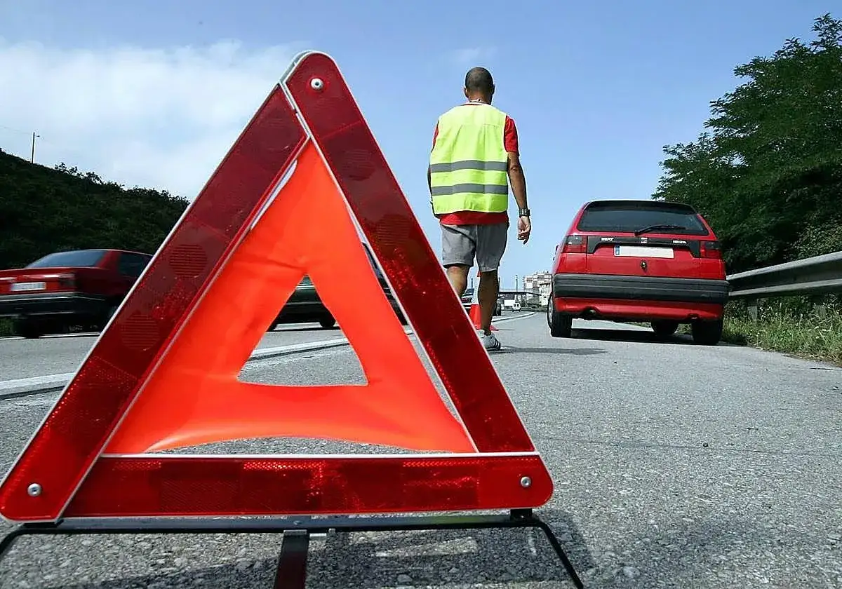 Triángulo de emergencia en primer plano. Un hombre con chaleco reflectante se aleja de un coche rojo averiado. Hay que avisar pas.