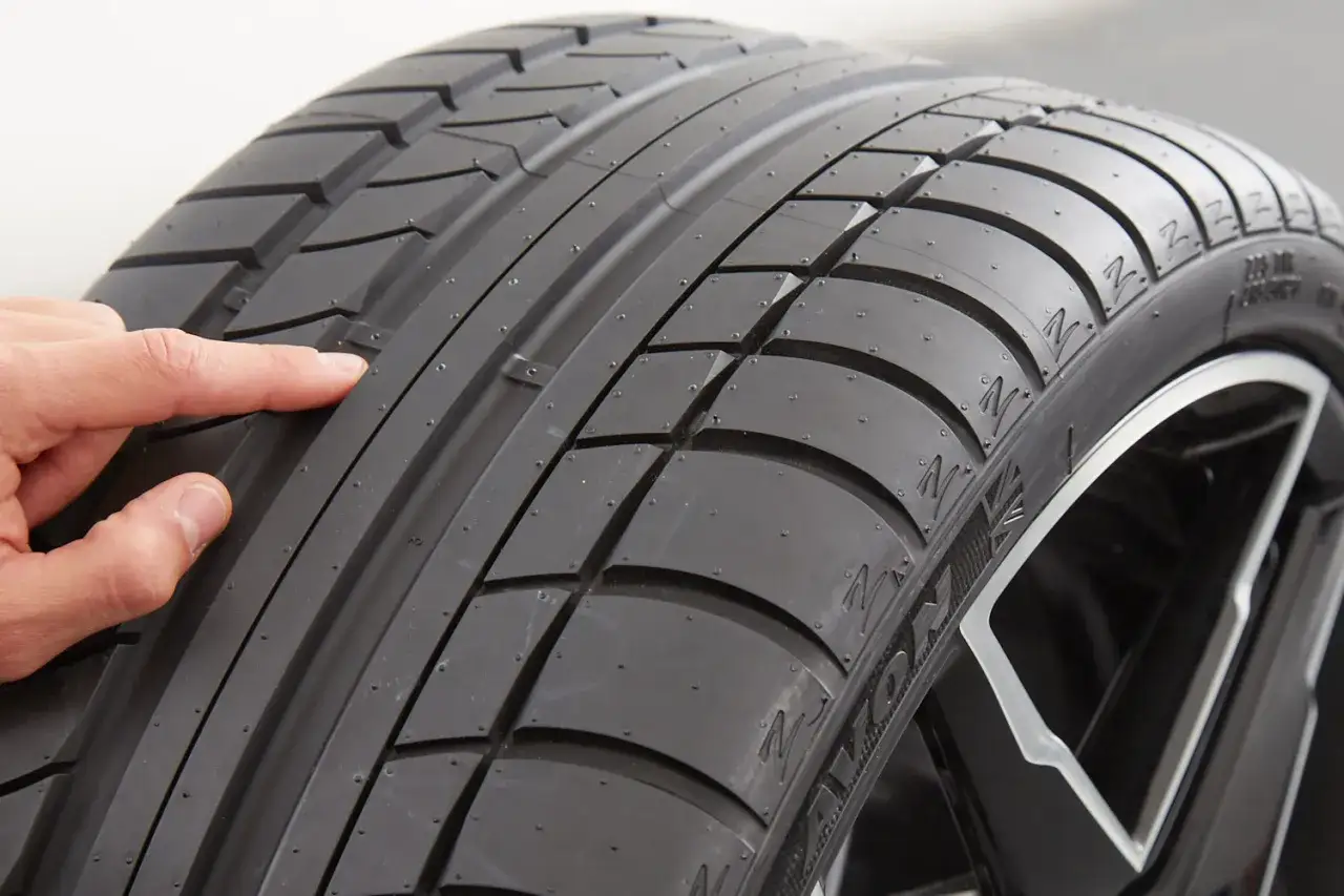 Un dedo se&ntilde;ala la banda de rodadura de un neum&aacute;tico, mostrando su dise&ntilde;o y textura. Estos son los neum&aacute;ticos, esenciales para la tracci&oacute;n y seguridad del veh&iacute;culo.