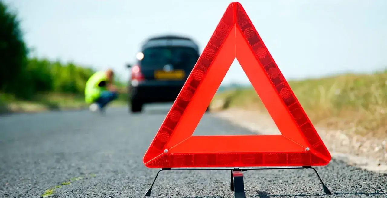 Triángulo de emergencia rojo en la carretera. Al fondo, una persona con chaleco reflectante y un coche averiado. Esto es lo que significa