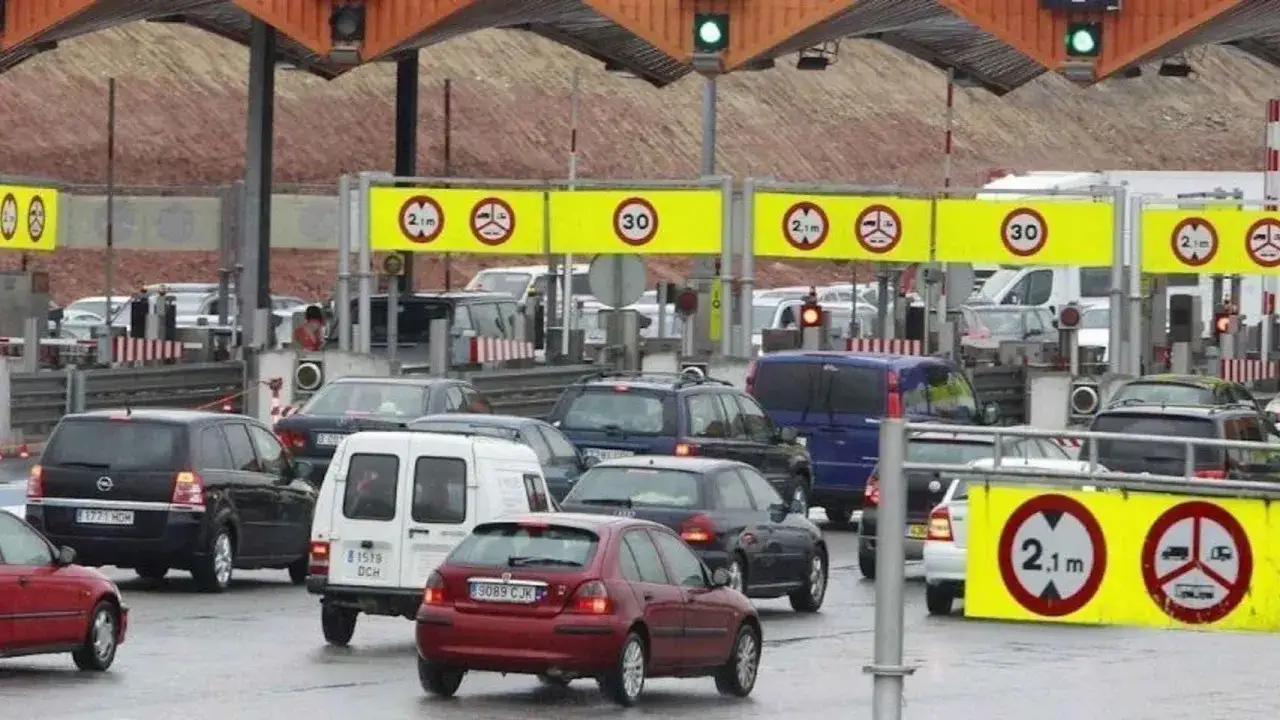 Tráfico en la autopista Pau Casals, con coches esperando en las cabinas de peaje. Señales de tráfico indican restricciones de altura y velocidad.