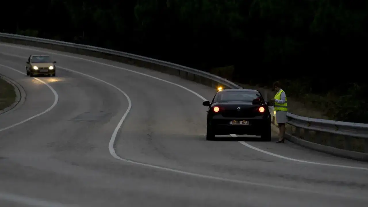 Una mujer con chaleco reflectante junto a un coche negro con luces de emergencia, indicando un posible accidente por objeto en la carretera.