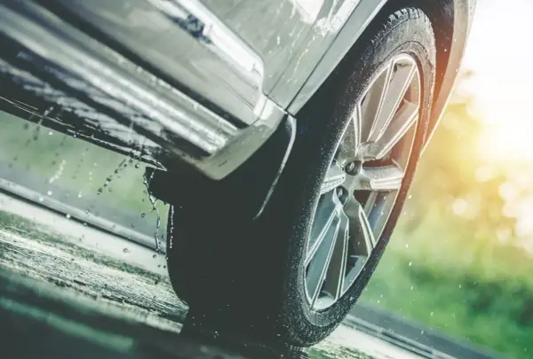 Rueda de coche sobre asfalto mojado, con gotas de agua salpicando, indicando una posible **pérdida de adherencia** en condiciones de lluvia.